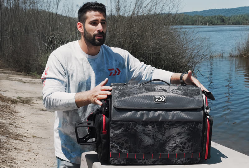 A man holding a Daiwa Tackle Barn fishing tackle bag by the water