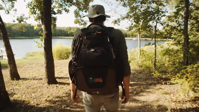 A man with a Wild River Tackle Tek Nomad Lighted Backpack designed for fishing gear, walking through the woods